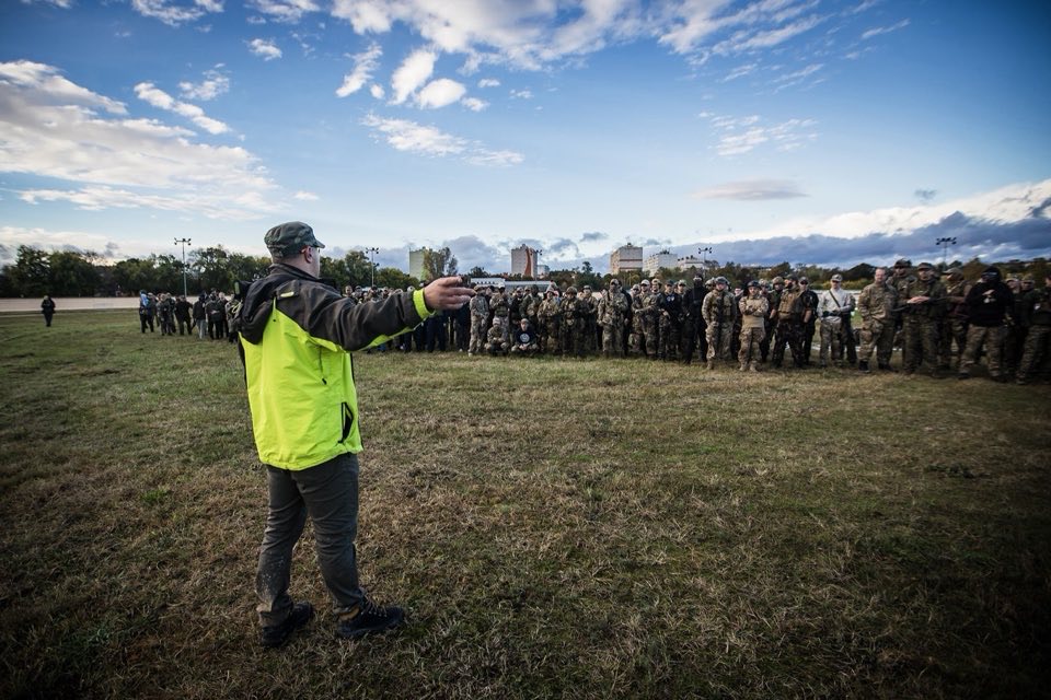 A staff member in a high-visibility vest addressing a large group of participants on a field.