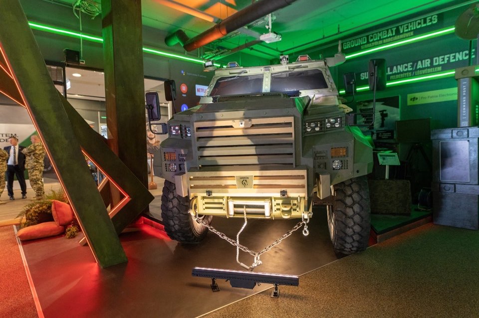 An armoured truck on display under green and red lighting, framed by timber structures.