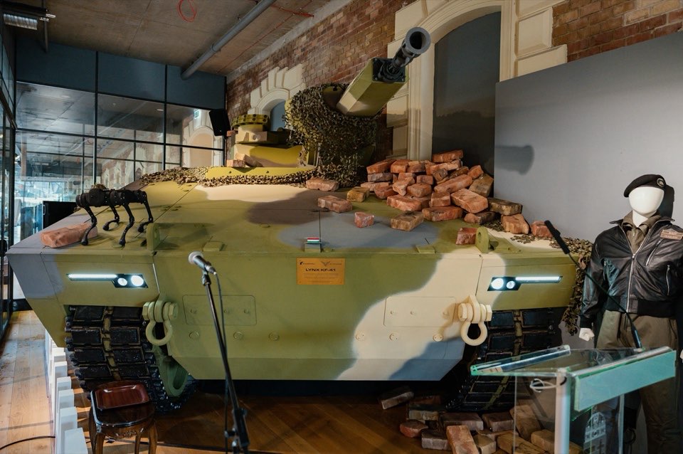 An armoured vehicle in camouflage paint displayed indoors beside a uniformed mannequin.
