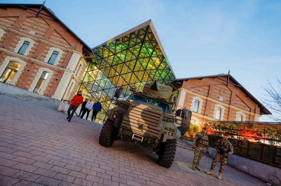 A military vehicle on display in front of a glass-fronted building, with visitors in the square.