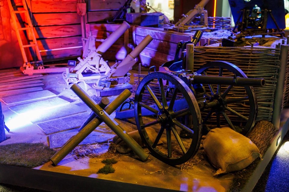 Exhibition scene with a historic field gun, sandbags and anti-tank obstacles under coloured lighting.