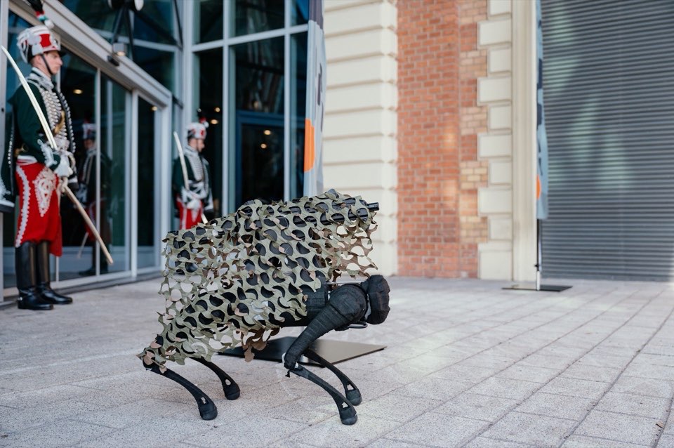 A four-legged robot draped in camouflage netting on the pavement outside a building.