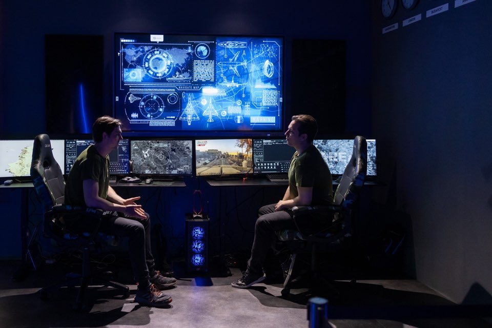 Two people seated in front of a wall of monitors in a darkened simulation room.