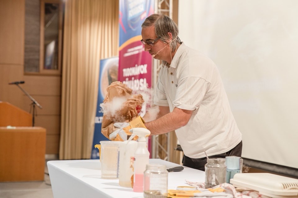 A presenter demonstrating a chemistry experiment with flasks at a table on stage.