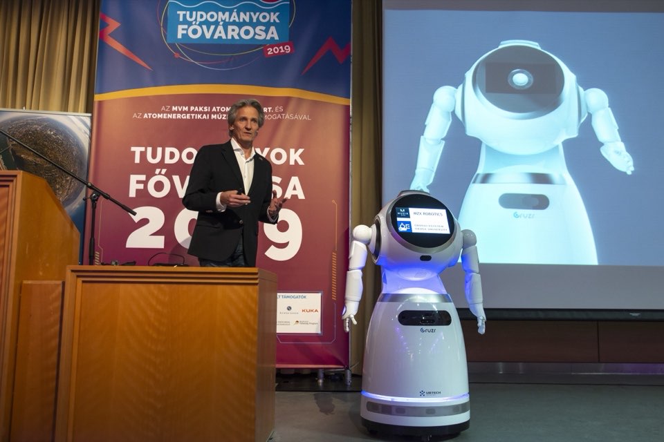 A speaker at a lectern beside a white humanoid service robot, with a robot image on the screen behind.