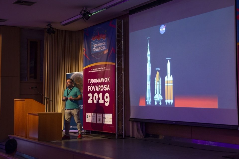 A speaker on a dark stage beside a projection of launch rockets.