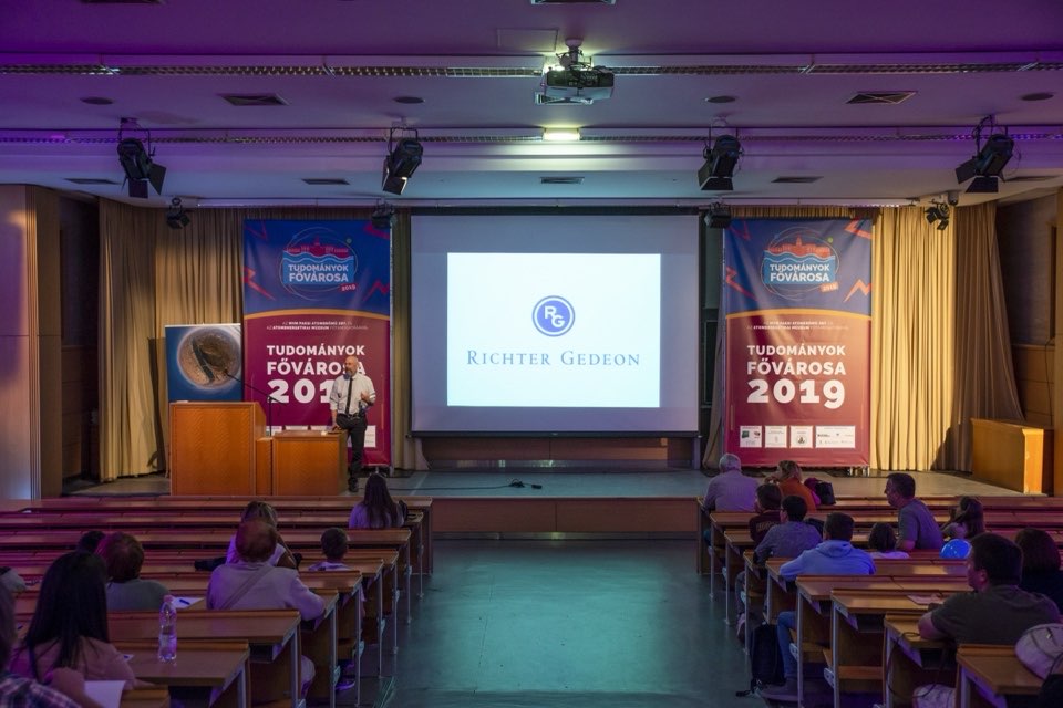 A wide view of a lecture hall with festival banners either side of a large projection screen.