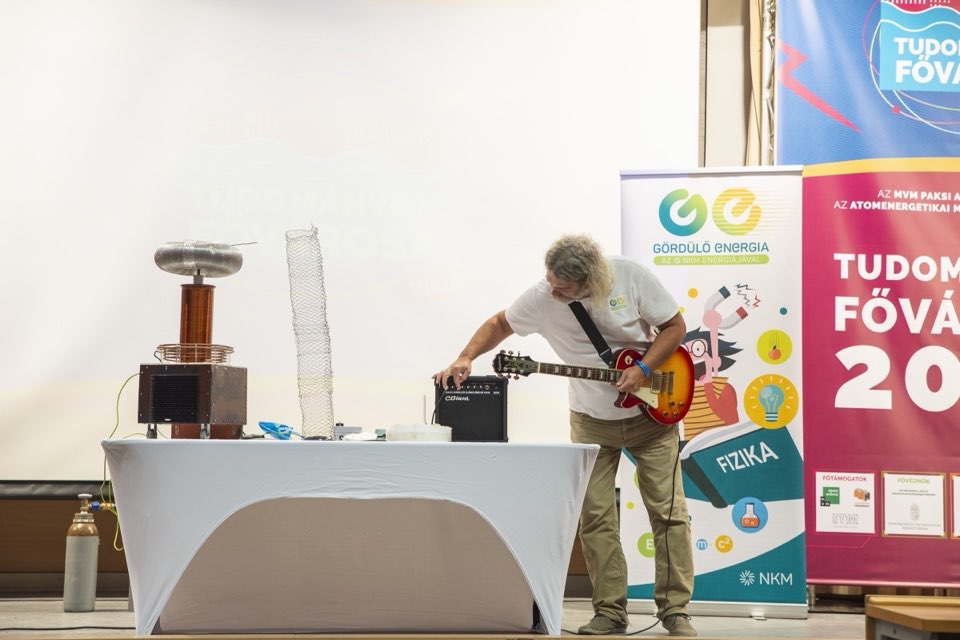A performer playing an electric guitar beside a Tesla coil on a stage with festival banners.