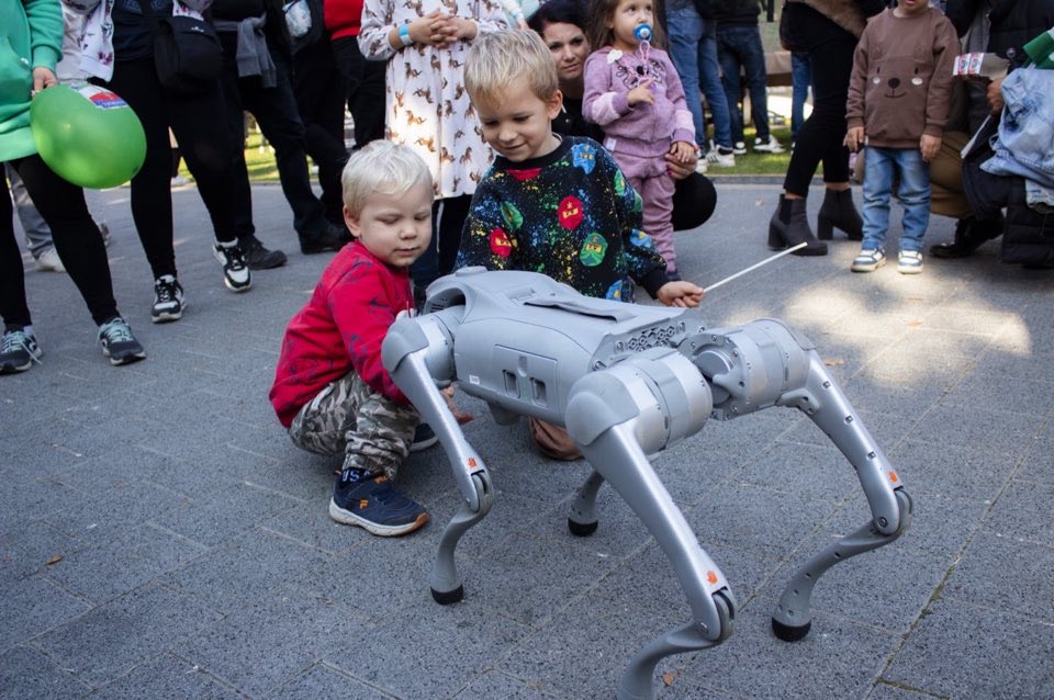 Children watching a small four-legged robot at a busy outdoor family event.