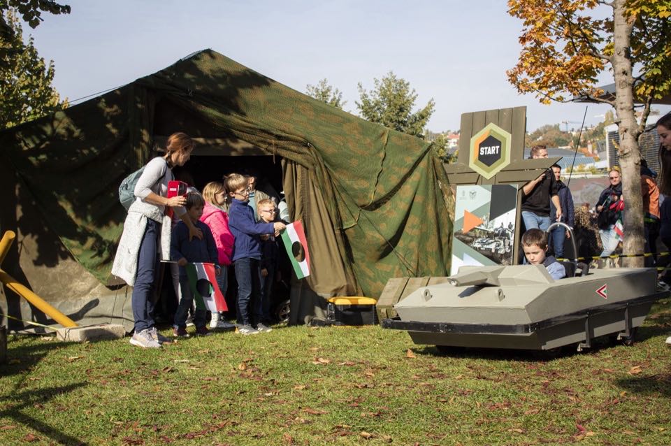 Families gathered beside a military tent and a vehicle at an outdoor event.