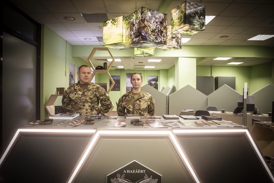 Two uniformed staff members behind an illuminated reception desk in a green-walled room.