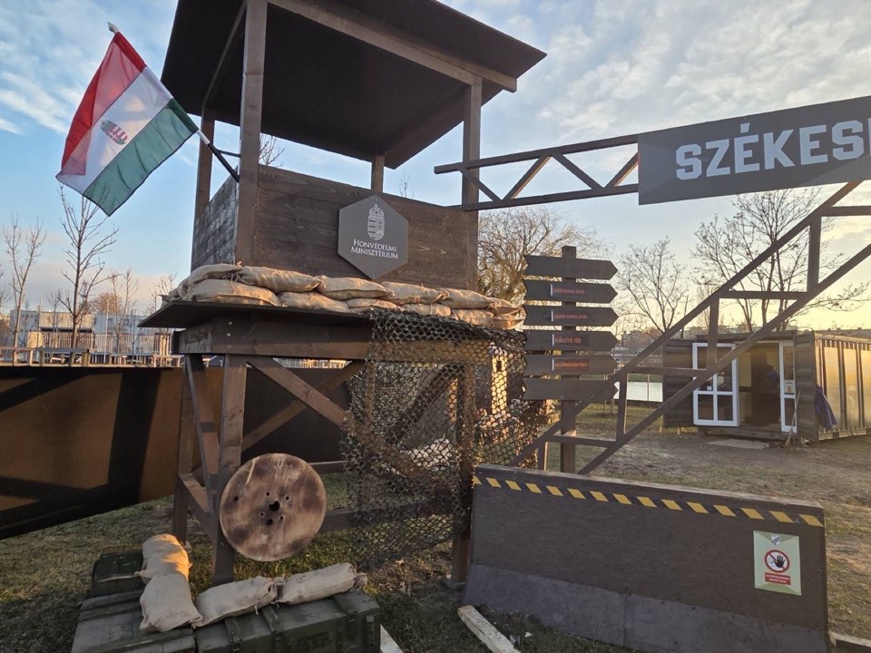 A timber watchtower with a Hungarian flag at the entrance to an outdoor venue.