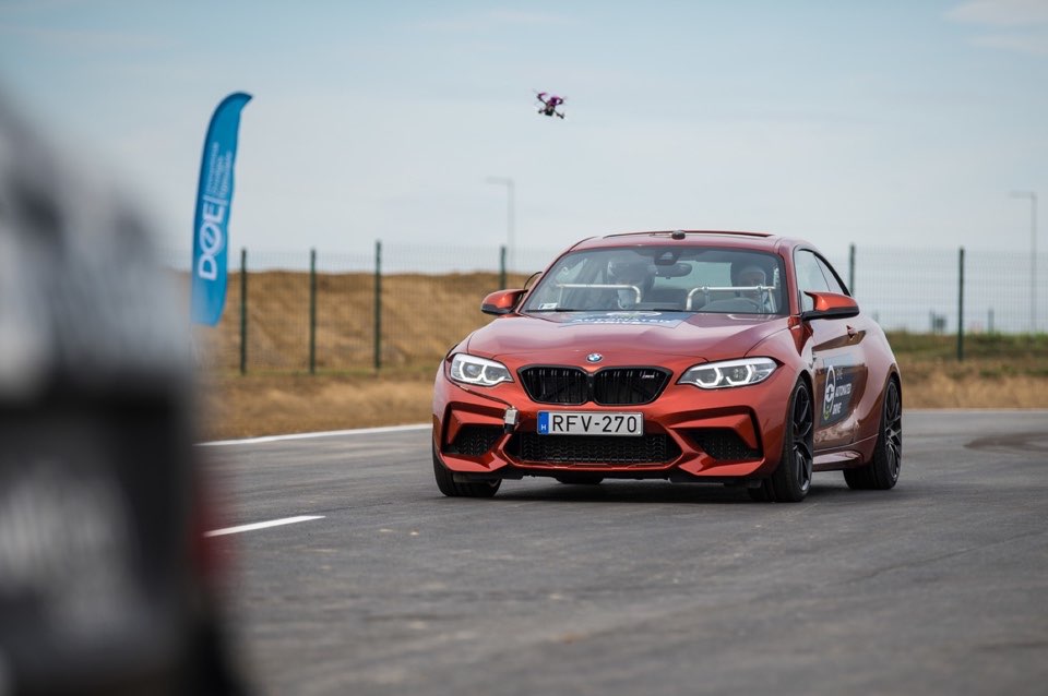 A car on a test track, with a camera drone in the air beside it.